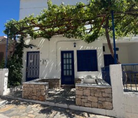 Dom z winoroślą nad drzwiami – Skopelos Biały dom z niebieskimi drzwiami i winoroślą tworzącą naturalną pergolę na Skopelos